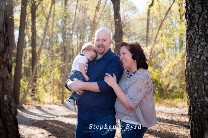 A father holds son while mom laughs during an outdoor family photo session in the woods at Lake Benson Park.