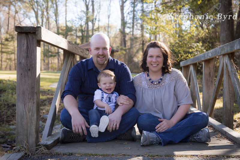 Two parents sit outside on a wooden bridge with their one year old boy enjoying the sun light.